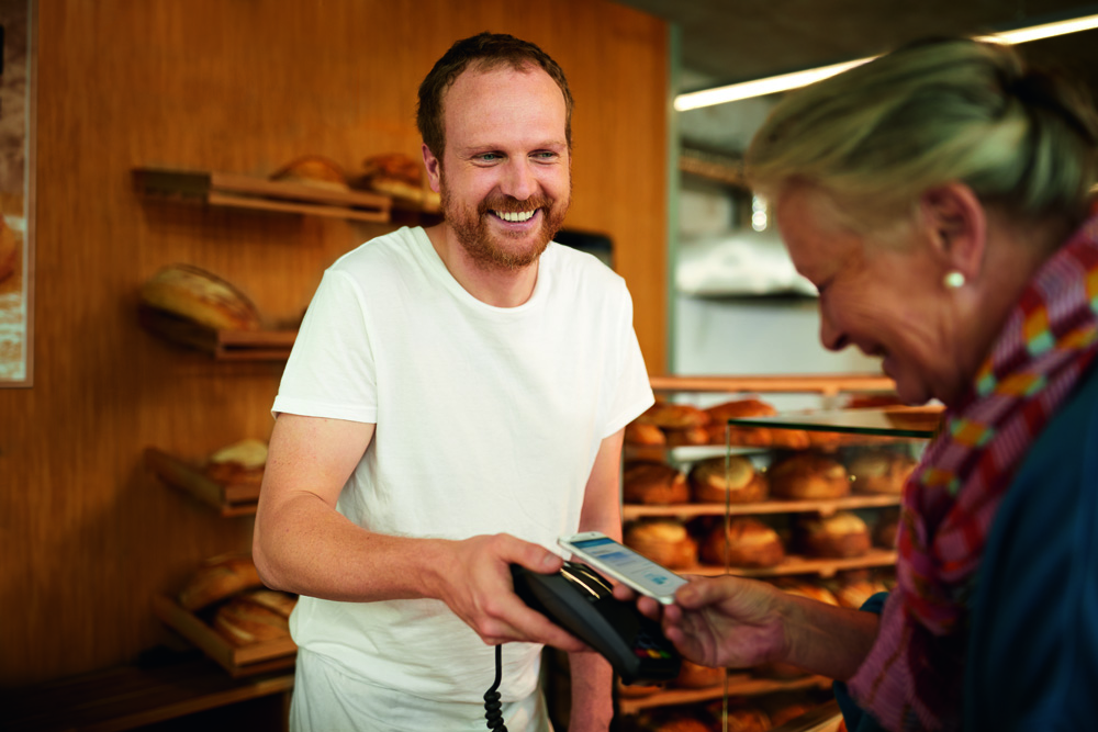 Ein Bäcker in weißer Kleidung lächelt freundlich, während er in einer Bäckerei ein Kartenterminal hält. Eine Kundin bezahlt kontaktlos mit ihrem Smartphone. Im Hintergrund sind Regale mit verschiedenen Broten zu sehen. Die Szene vermittelt eine freundliche und moderne Einkaufsatmosphäre. Ein Bäcker in weißer Kleidung lächelt freundlich, während er in einer Bäckerei ein Kartenterminal hält. Eine Kundin bezahlt kontaktlos mit ihrem Smartphone. Im Hintergrund sind Regale mit verschiedenen Broten zu sehen. Die Szene vermittelt eine freundliche und moderne Einkaufsatmosphäre.