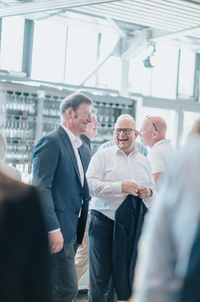 Mehrere Männer in Hemden und Anzügen stehen in einer hellen, modernen Umgebung mit großen Fenstern und unterhalten sich angeregt und lachen. Die Atmosphäre wirkt geschäftlich und freundlich. Mehrere Männer in Hemden und Anzügen stehen in einer hellen, modernen Umgebung mit großen Fenstern und unterhalten sich angeregt und lachen. Die Atmosphäre wirkt geschäftlich und freundlich.