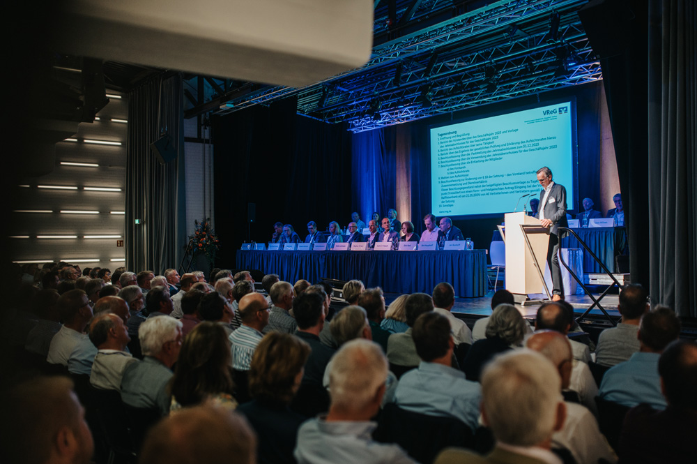 Ein großer Saal ist gefüllt mit zahlreichen Zuhörern, die aufmerksam einer Rede folgen, die von einem Mann an einem Rednerpult auf der Bühne gehalten wird. Hinter dem Redner sitzt eine Gruppe weiterer Personen an einem langen Tisch vor einer großen Leinwand mit Text und Logos. Die Beleuchtung ist auf die Bühne gerichtet, während der Zuschauerbereich etwas dunkler ist. Ein großer Saal ist gefüllt mit zahlreichen Zuhörern, die aufmerksam einer Rede folgen, die von einem Mann an einem Rednerpult auf der Bühne gehalten wird. Hinter dem Redner sitzt eine Gruppe weiterer Personen an einem langen Tisch vor einer großen Leinwand mit Text und Logos. Die Beleuchtung ist auf die Bühne gerichtet, während der Zuschauerbereich etwas dunkler ist.
