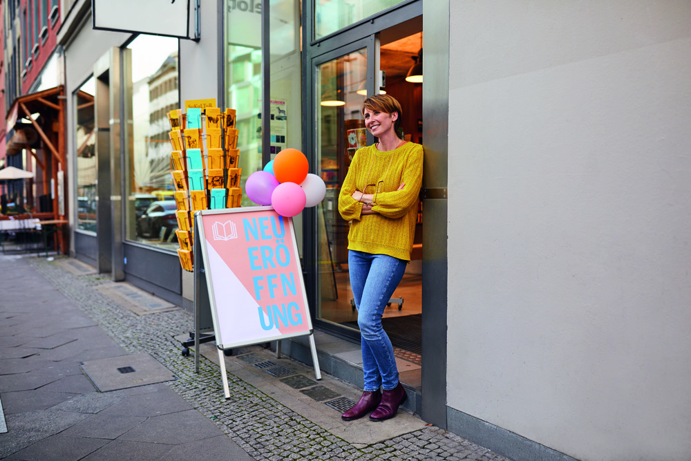 Eine lächelnde Frau in gelbem Pullover lehnt vor einem Ladenlokal mit einem "NEU ERÖFFNUNG"-Aufsteller und Luftballons. Eine lächelnde Frau in gelbem Pullover lehnt vor einem Ladenlokal mit einem "NEU ERÖFFNUNG"-Aufsteller und Luftballons.
