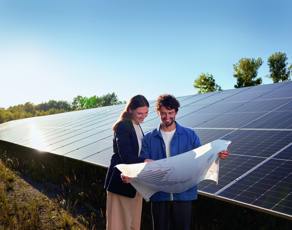 Zwei Personen betrachten gemeinsam einen großen Plan vor einer Reihe von Solarmodulen unter einem blauen Himmel. Zwei Personen betrachten gemeinsam einen großen Plan vor einer Reihe von Solarmodulen unter einem blauen Himmel.