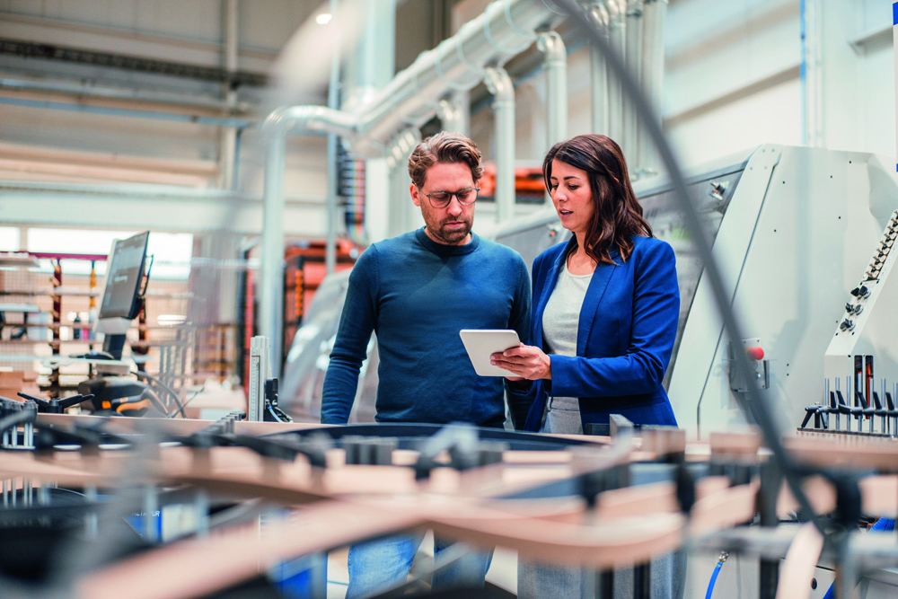 Ein Mann mit Brille und eine Frau im Blazer betrachten ein Tablet in einer Produktionshalle mit Maschinen. Ein Mann mit Brille und eine Frau im Blazer betrachten ein Tablet in einer Produktionshalle mit Maschinen.