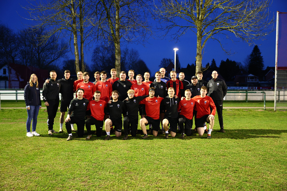 Eine große Gruppe von Fußballspielern in roten und schwarzen Trikots sowie einige Betreuer in schwarzen Trainingsanzügen posieren für ein Mannschaftsfoto auf einem grünen Fußballplatz in der Abenddämmerung. Im Hintergrund sind Bäume, Häuser und eine Straßenlaterne unter einem dunkler werdenden Himmel zu sehen.