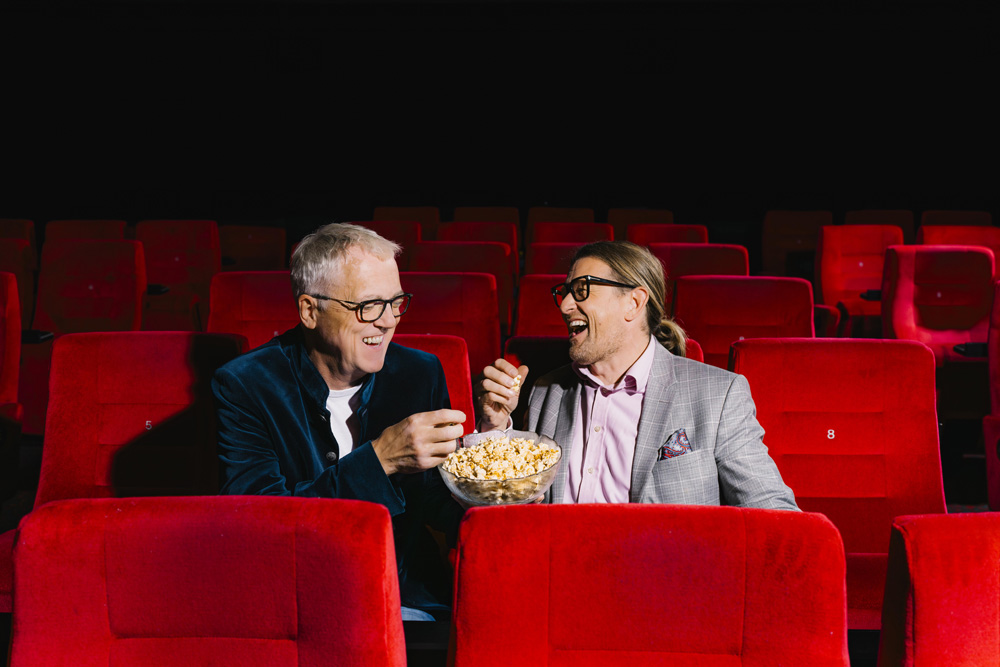Zwei Männer sitzen in einem Kinosaal mit roten Sitzen. Sie lachen und teilen sich eine Schüssel Popcorn.
