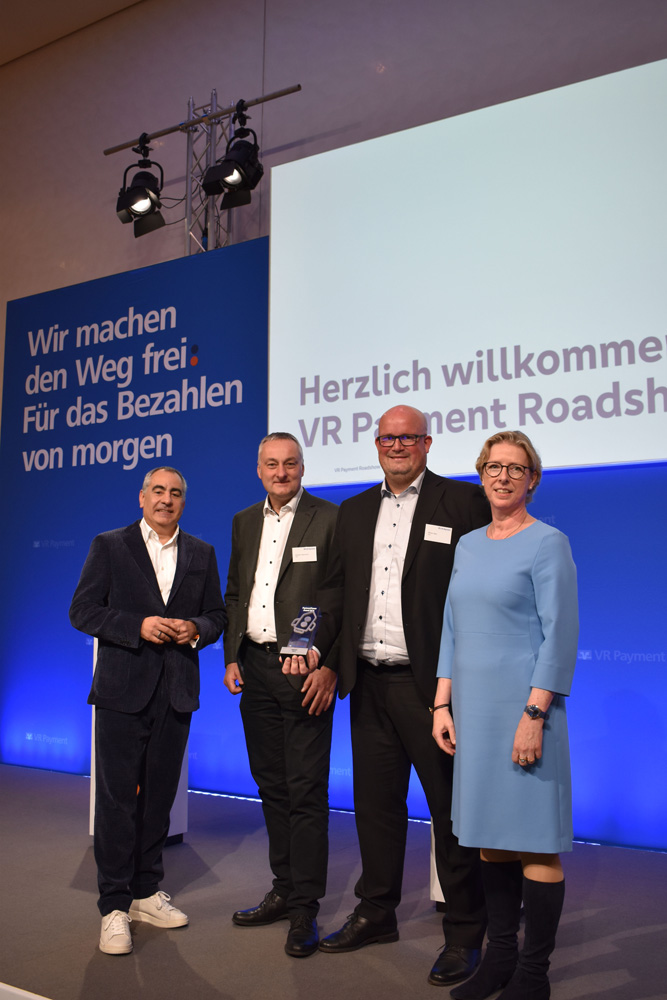 Vier Personen stehen auf einer Bühne vor einer blauen Wand mit weißer Aufschrift, darunter "Wir machen den Weg frei. Für das Bezahlen von morgen" und "Herzlich willkommen VR Payment Roadshow". Einer der Männer hält eine Auszeichnung. Die Beleuchtung ist hell und auf die Gruppe gerichtet. Vier Personen stehen auf einer Bühne vor einer blauen Wand mit weißer Aufschrift, darunter "Wir machen den Weg frei. Für das Bezahlen von morgen" und "Herzlich willkommen VR Payment Roadshow". Einer der Männer hält eine Auszeichnung. Die Beleuchtung ist hell und auf die Gruppe gerichtet.