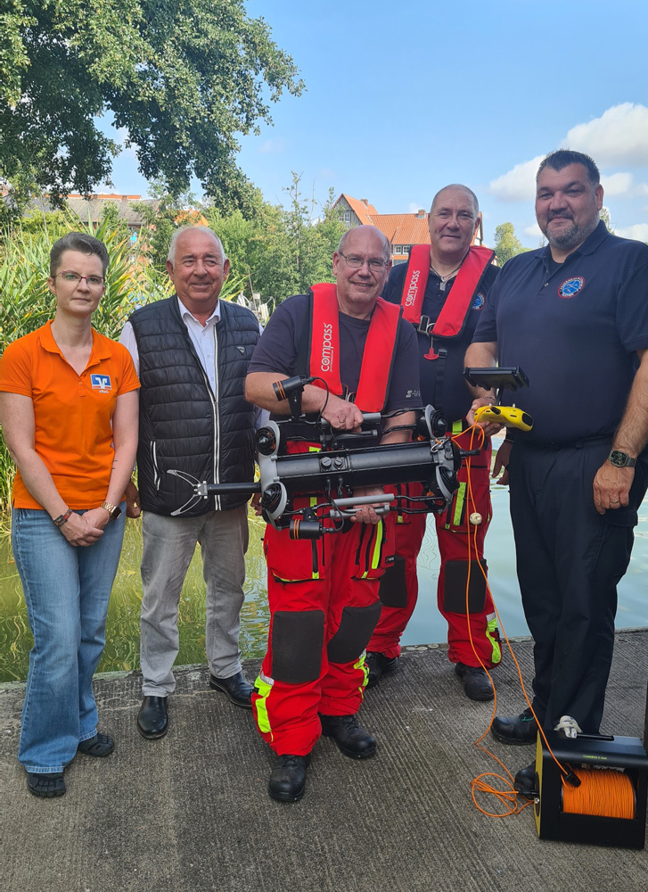 Fünf Personen stehen im Freien neben einem Gewässer. Drei der Männer tragen rote Rettungsanzüge und Schwimmwesten und halten Tauchausrüstung, darunter einen Unterwasserroboter. Die Frau links trägt ein orangefarbenes Poloshirt mit einem Banklogo und Jeans, der Mann neben ihr eine dunkle Weste über einem hellen Hemd. Der Mann rechts trägt ein dunkelblaues Poloshirt. Im Hintergrund sind Bäume und Gebäude unter einem leicht bewölkten Himmel zu sehen.