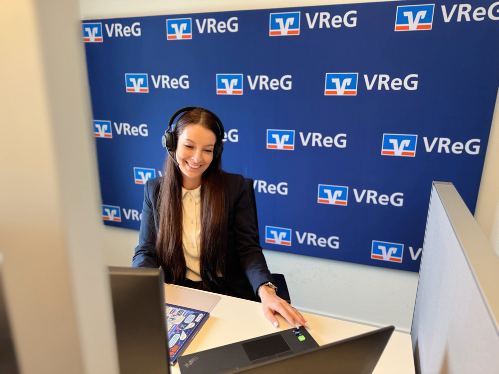 ine junge Frau mit langen, braunen Haaren und einem Headset sitzt lächelnd an einem Schreibtisch und arbeitet an einem Laptop. Hinter ihr ist eine blaue Wand mit dem weißen "VReG"-Logo mehrmals wiederholt zu sehen. Die Beleuchtung ist hell und gleichmäßig. Eine junge Frau mit langen, braunen Haaren und einem Headset sitzt lächelnd an einem Schreibtisch und arbeitet an einem Laptop. Hinter ihr ist eine blaue Wand mit dem weißen "VReG"-Logo mehrmals wiederholt zu sehen. Die Beleuchtung ist hell und gleichmäßig.