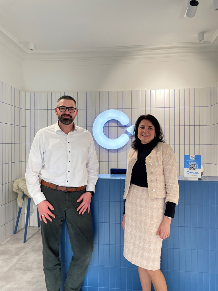 Zwei Personen, ein Mann mit Bart und Brille in einem weißen Hemd und eine Frau in einem hellen Tweed-Kostüm, stehen lächelnd vor einem Empfangsbereich mit einer blauen Theke und einem beleuchteten "C"-Logo an einer weiß gefliesten Wand.