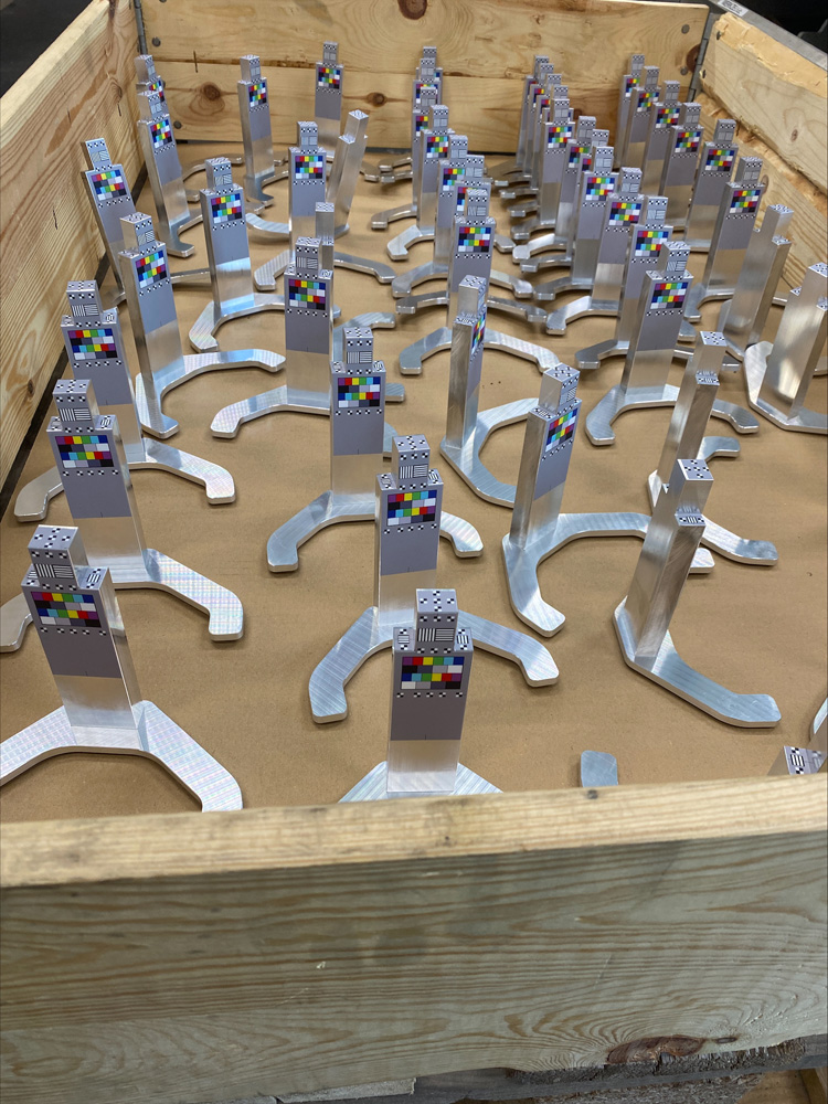 Ein Holzkasten ist gefüllt mit zahlreichen identischen, silberfarbenen Metallteilen, die jeweils mit einem kleinen, bunten Testmusteraufkleber versehen sind. Ein Holzkasten ist gefüllt mit zahlreichen identischen, silberfarbenen Metallteilen, die jeweils mit einem kleinen, bunten Testmusteraufkleber versehen sind.