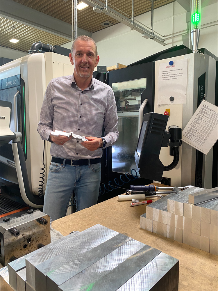Ein Mann mittleren Alters steht lächelnd in einer Werkshalle vor einer CNC-Maschine und hält ein kleines Metallteil in den Händen. Auf einem Tisch vor ihm liegen weitere Werkstücke und Werkzeuge.