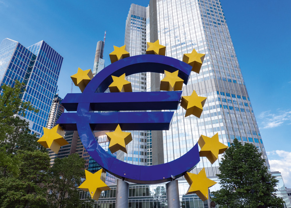 Ein großes, blaues Euro-Symbol, umgeben von gelben Sternen, steht vor der Skyline von Frankfurt am Main. Im Hintergrund ragen moderne Wolkenkratzer in den blauen Himmel mit einigen weißen Wolken. Das Euro-Zeichen und die Sterne sind auf Metallpfosten montiert und von Bäumen umgeben. Ein großes, blaues Euro-Symbol, umgeben von gelben Sternen, steht vor der Skyline von Frankfurt am Main. Im Hintergrund ragen moderne Wolkenkratzer in den blauen Himmel mit einigen weißen Wolken. Das Euro-Zeichen und die Sterne sind auf Metallpfosten montiert und von Bäumen umgeben.