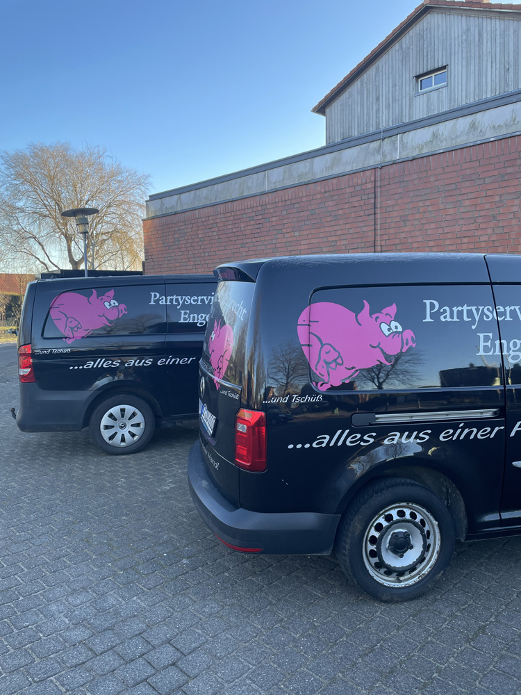 Zwei schwarze Lieferwagen mit pinkfarbenem Schweine-Logo und der Aufschrift "Partyservice Engelbrecht" sowie "...alles aus einer Hand" parken auf einem gepflasterten Platz vor einem grauen Gebäude unter einem klaren, blauen Himmel. Zwei schwarze Lieferwagen mit pinkfarbenem Schweine-Logo und der Aufschrift "Partyservice Engelbrecht" sowie "...alles aus einer Hand" parken auf einem gepflasterten Platz vor einem grauen Gebäude unter einem klaren, blauen Himmel.