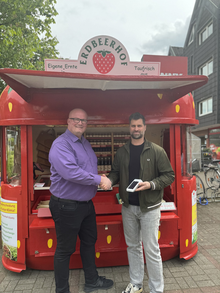 Zwei Männer schütteln sich vor einem roten Verkaufsstand in Form einer Erdbeere mit der Aufschrift "Erdbeerhof". Der Mann links trägt ein lila Hemd und eine Brille, der Mann rechts eine grüne Jacke und hält ein weißes Kartenterminal in der Hand. Der Stand bietet "Eigene Ernte" und "Taufrisch aus..." an. Im Hintergrund sind Gebäude und Bäume zu sehen.