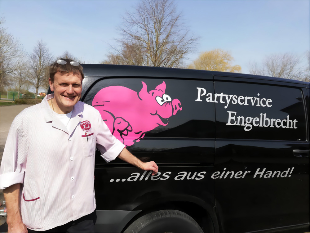 Ein lächelnder Mann in einer weißen Kochjacke mit roten Akzenten steht neben einem schwarzen Lieferwagen, der mit einem großen pinkfarbenen Schwein, der Aufschrift "Partyservice Engelbrecht" und dem Slogan "...alles aus einer Hand!" bedruckt ist. Der Hintergrund zeigt einen Parkplatz und Bäume unter einem hellen Himmel.