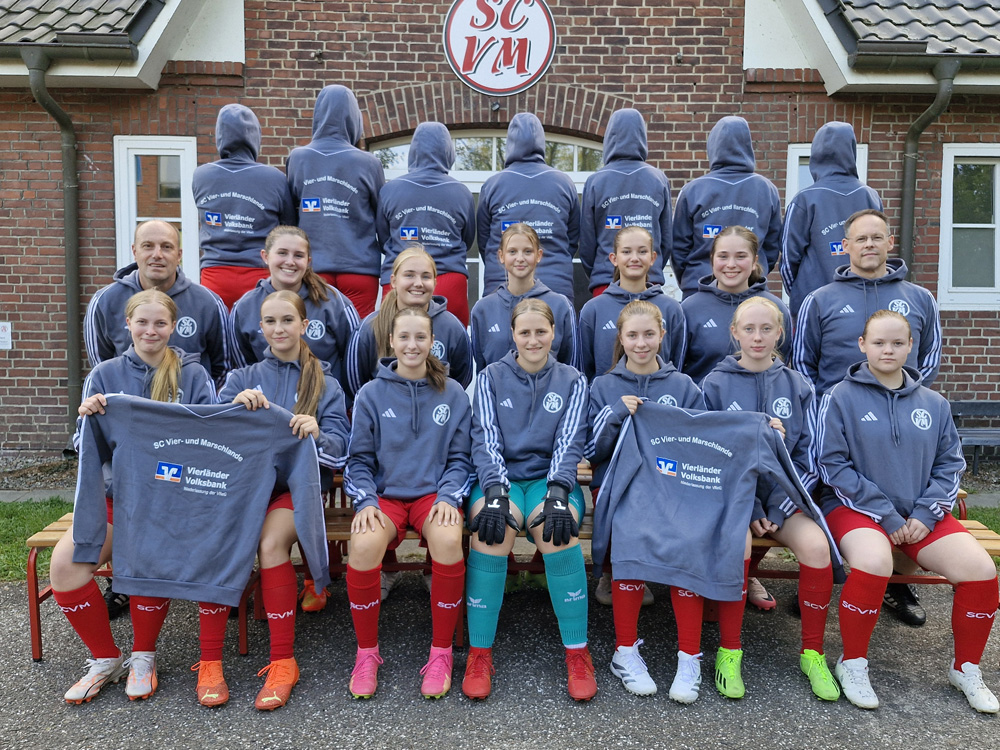 Eine Jugendmannschaft des SC Vier- und Marschlande posiert für ein Gruppenfoto vor einem Backsteingebäude. Die Spielerinnen und Trainer tragen graue Kapuzenpullover mit Vereinslogo und Sponsorenaufdruck sowie rote Hosen und Stutzen in unterschiedlichen Farbkombinationen. Einige Spielerinnen halten zusätzliche Pullover mit dem Vereinsnamen und Sponsorenlogo in die Kamera.