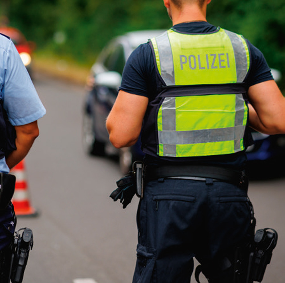Zwei Polizisten in blauer Uniform und neongelben Sicherheitswesten mit der Aufschrift "POLIZEI" stehen auf einer Straße. Im Hintergrund sind unscharf ein Auto und Verkehrskegel zu erkennen. Zwei Polizisten in blauer Uniform und neongelben Sicherheitswesten mit der Aufschrift "POLIZEI" stehen auf einer Straße. Im Hintergrund sind unscharf ein Auto und Verkehrskegel zu erkennen.