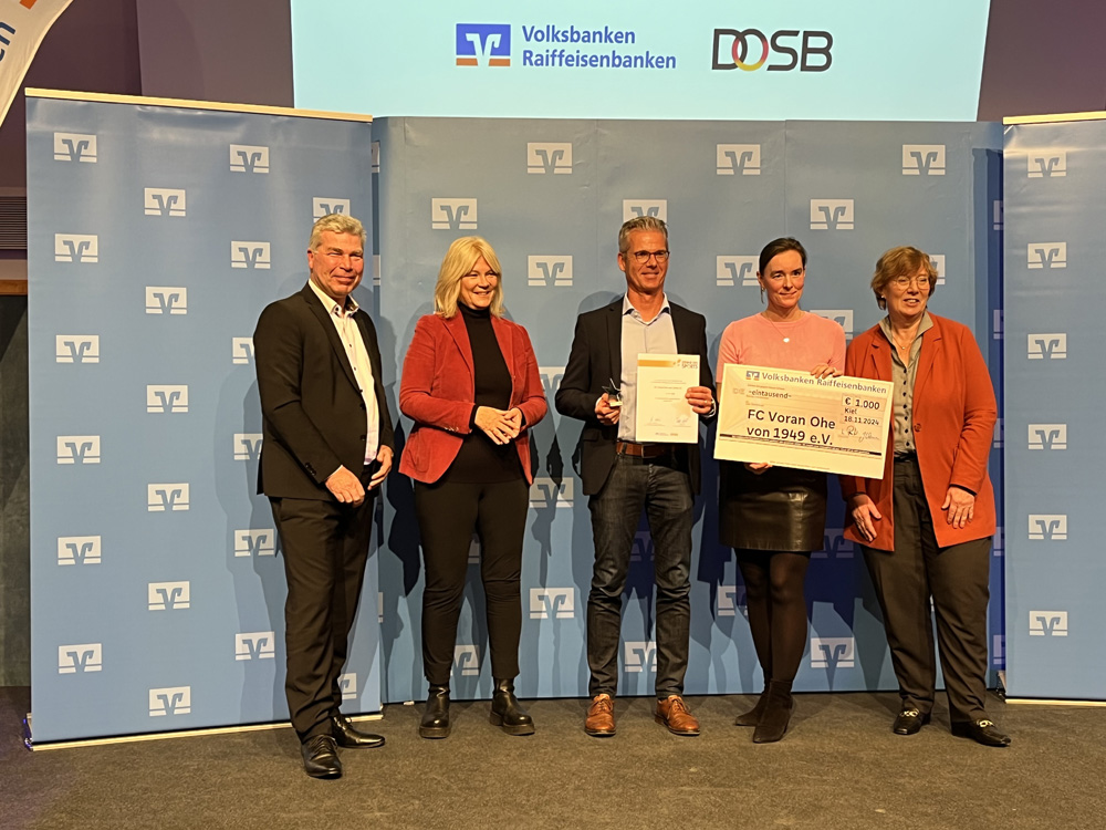 Fünf Personen stehen auf einer Bühne vor einer blauen Wand mit dem Logo der Volksbanken Raiffeisenbanken und des DOSB. Die mittlere Person hält eine Urkunde und einen überdimensionalen Scheck über 1.000 Euro für den "FC Voran Ohe von 1949 e.V.". Die anderen vier Personen lächeln in die Kamera.