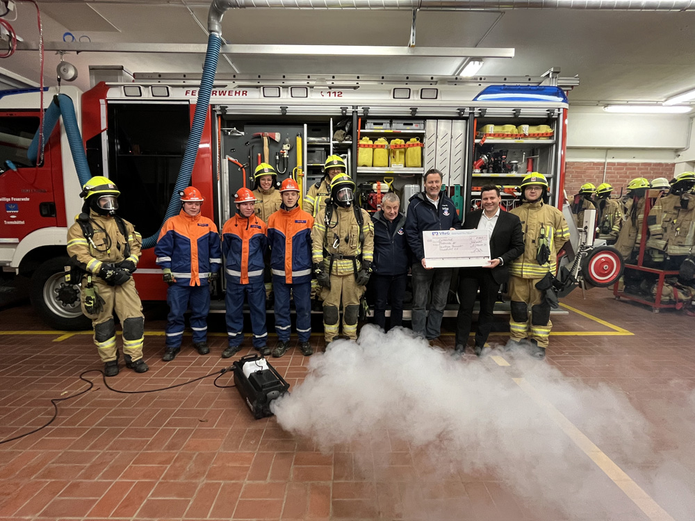 Eine Gruppe von Feuerwehrleuten in Einsatzkleidung, einige mit Atemschutzmasken, und zwei zivile Personen stehen in einer Feuerwehrgarage vor einem roten Feuerwehrfahrzeug. Eine der zivilen Personen überreicht der Gruppe einen großen Scheck. Im Vordergrund ist eine Nebelmaschine aktiv und erzeugt weißen Rauch.