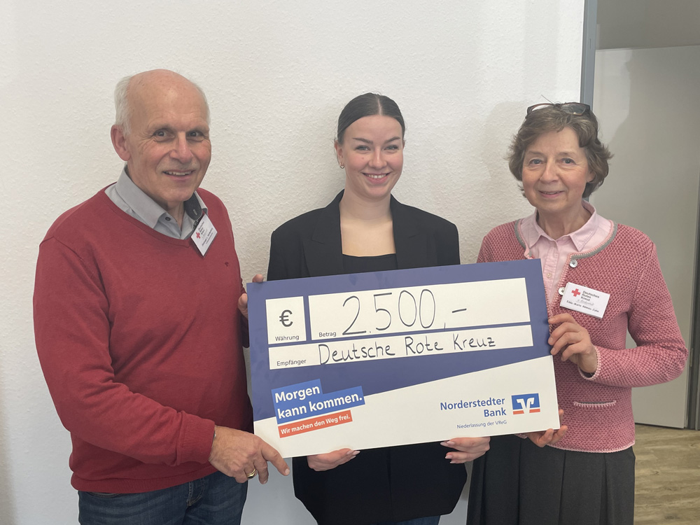 Drei Personen stehen lächelnd vor einer hellen Wand und halten einen großen symbolischen Scheck über 2500 Euro, ausgestellt von der Norderstedter Bank für das Deutsche Rote Kreuz. Die beiden älteren Personen tragen Kleidung mit dem Roten Kreuz Symbol.