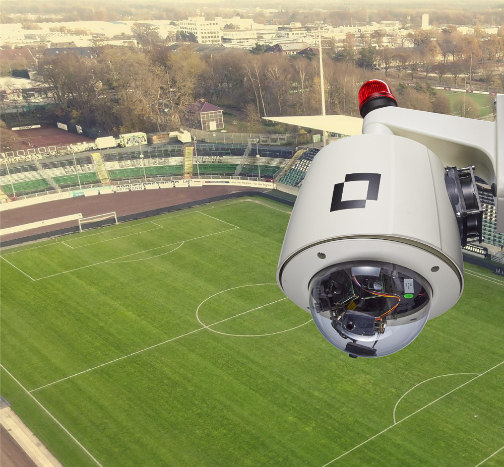 Eine hochmoderne, weiße Überwachungskamera mit einem roten Warnlicht ist in erhöhter Position angebracht und überblickt ein grünes Fußballstadion mit weißen Linien und leeren Tribünen. Im Hintergrund erstreckt sich eine Stadtlandschaft unter einem trüben Himmel. Eine hochmoderne, weiße Überwachungskamera mit einem roten Warnlicht ist in erhöhter Position angebracht und überblickt ein grünes Fußballstadion mit weißen Linien und leeren Tribünen. Im Hintergrund erstreckt sich eine Stadtlandschaft unter einem trüben Himmel.