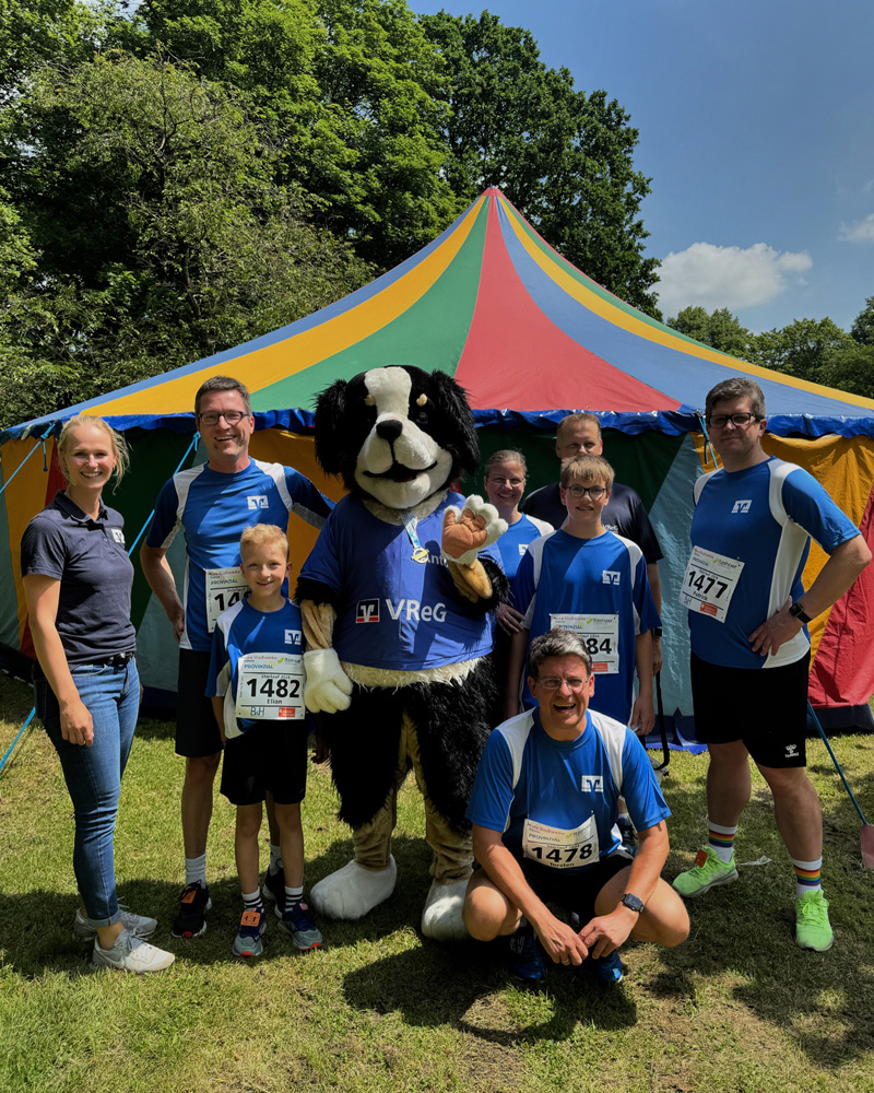 Eine Gruppe von Erwachsenen und Kindern in blauen Laufshirts mit Startnummern posiert mit einem Maskottchen in einem Hunde-Kostüm vor einem bunten Zirkuszelt im Freien. Die Stimmung wirkt fröhlich und gemeinschaftlich.