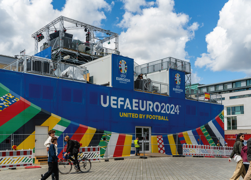 Ein großes, blaues Veranstaltungsgebäude mit der Aufschrift "UEFA EURO 2024 UNITED BY FOOTBALL" und dem dazugehörigen Logo steht auf einem gepflasterten Platz. Das Gebäude ist mit bunten, vertikalen Streifen in den Nationalfarben verschiedener europäischer Länder verziert. Menschen gehen und fahren Fahrräder davor. Der Himmel ist blau mit einigen weißen Wolken. Ein großes, blaues Veranstaltungsgebäude mit der Aufschrift "UEFA EURO 2024 UNITED BY FOOTBALL" und dem dazugehörigen Logo steht auf einem gepflasterten Platz. Das Gebäude ist mit bunten, vertikalen Streifen in den Nationalfarben verschiedener europäischer Länder verziert. Menschen gehen und fahren Fahrräder davor. Der Himmel ist blau mit einigen weißen Wolken.